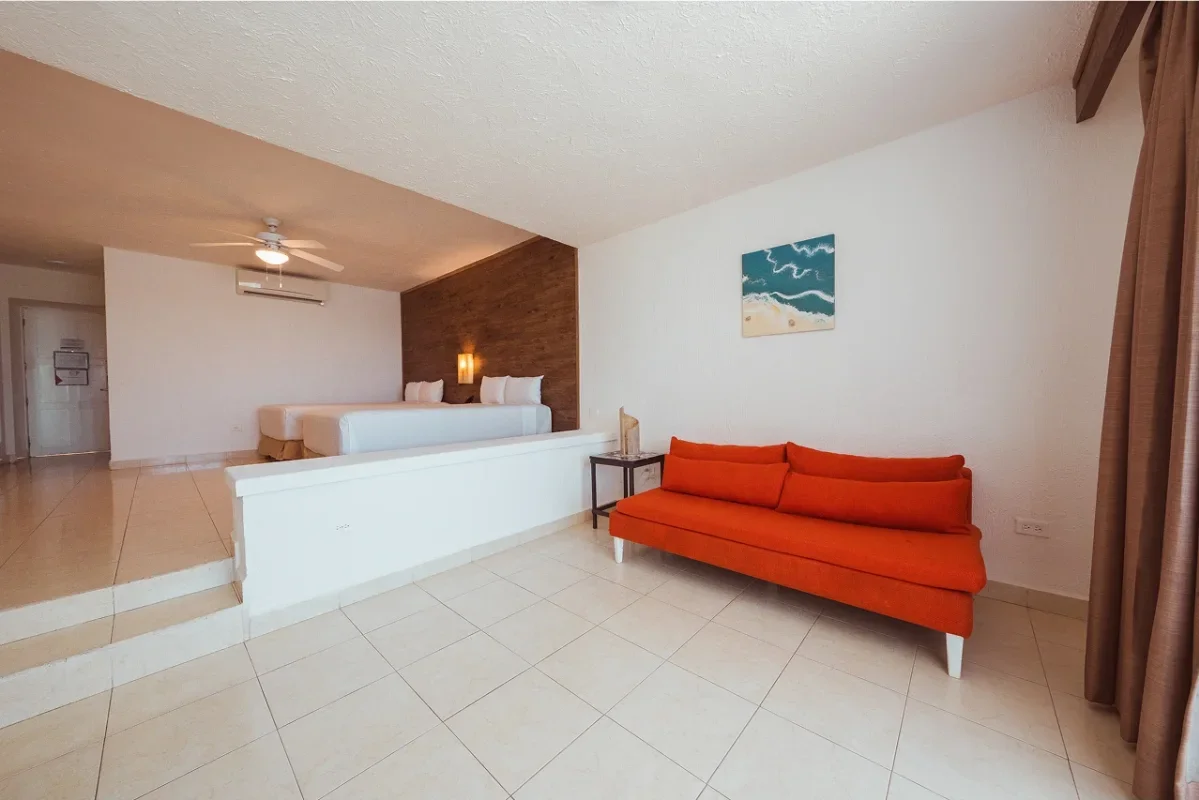 A hotel room with a small orange sofa, a side table, a beach-themed painting on the white wall, and a raised sleeping area with two beds against a wooden accent wall.