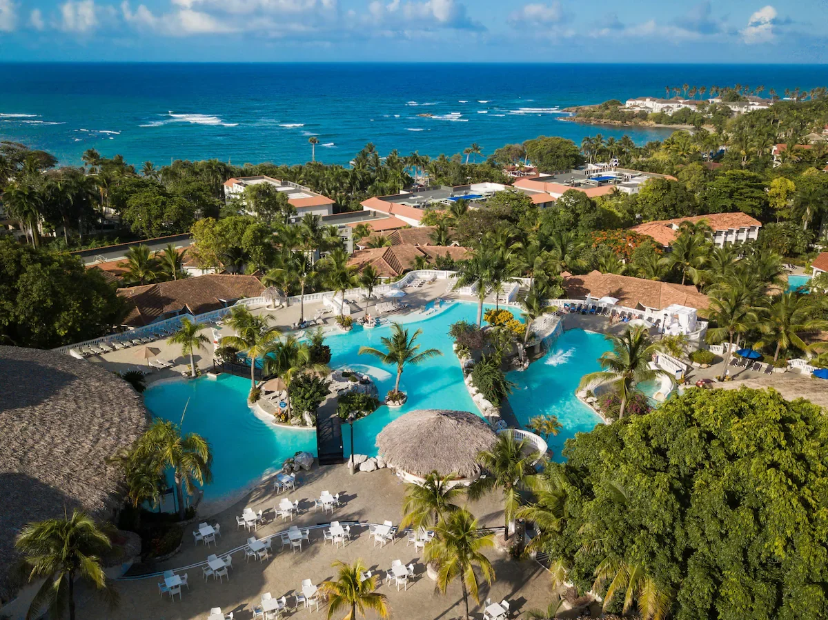 Aerial view of a tropical resort with swimming pools, surrounded by palm trees and lush greenery, with the ocean in the background.
