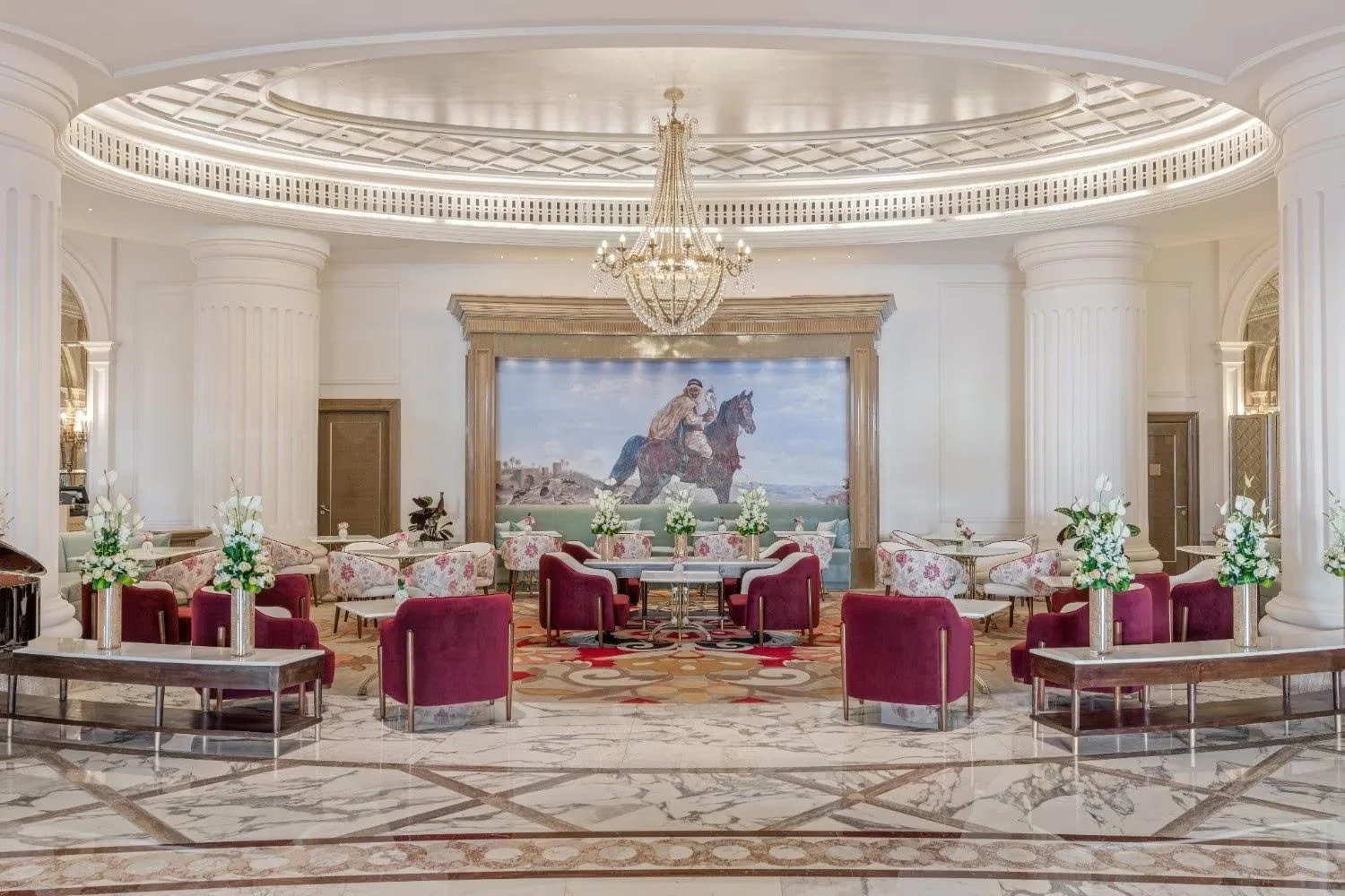 Elegant hotel lobby with white marble floors, purple velvet chairs, floral arrangements, a large painting of a person riding a horse, and a chandelier hanging from the ceiling.