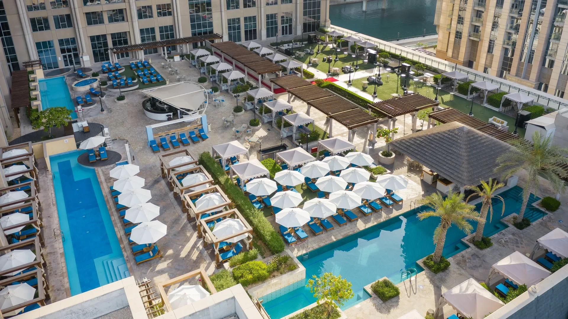 An aerial view of a luxury hotel rooftop pool area with multiple pools, sun loungers with umbrellas, cabanas, and a landscaped garden with trees, all adjacent to a body of water.
