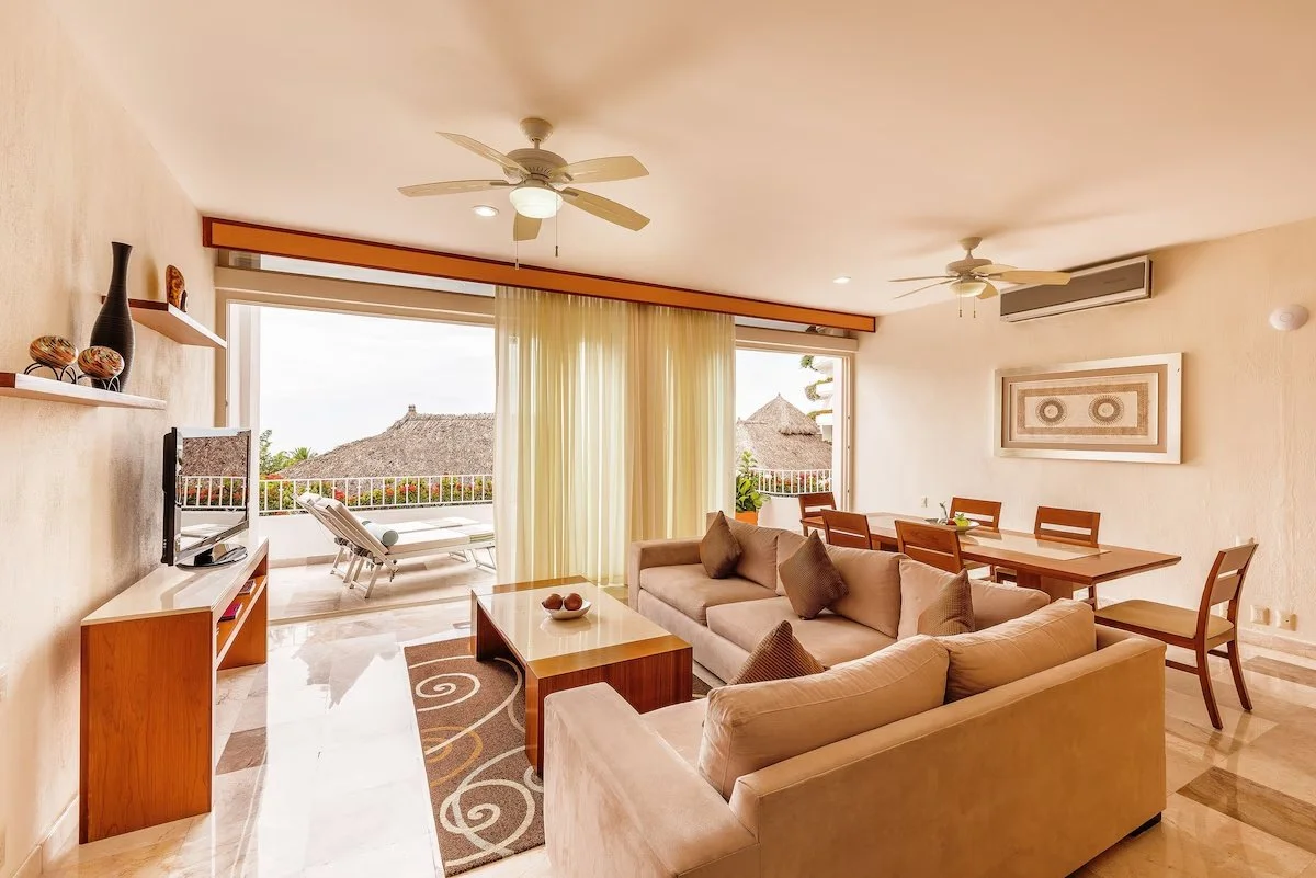 Living room with beige sofas, wooden dining table with chairs, sliding glass door with curtains, TV on a wooden stand, balcony with lounge chair and tropical view, ceiling fans, and wall art.