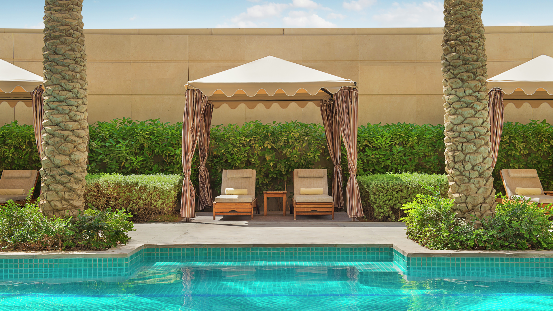 View of a swimming pool with turquoise tiles, beyond which there are outdoor lounge chairs under cabanas with striped curtains, surrounded by greenery and tall palm trees.