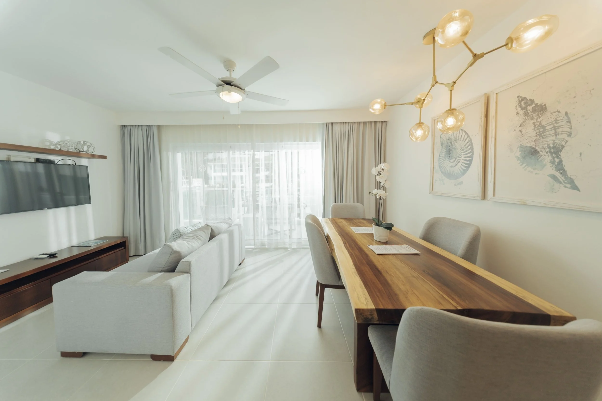 Living room with a ceiling fan, wall-mounted TV, white couch, wooden dining table with chairs, large window with curtains, and decorative wall art.