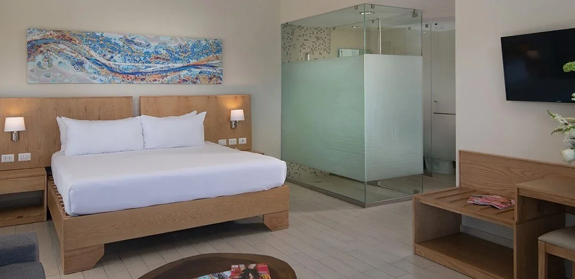 A modern hotel room featuring a large bed with white bedding, a wooden headboard, and wall-mounted lamps. There is a glass-enclosed bathroom with a shower, a flat-screen TV on the wall, and a wooden desk with a magazine and a chair. A colorful abstra