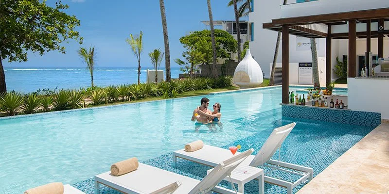 Couple swimming in a resort pool with poolside lounge chairs, beach, and tiki bar with drinks in the background