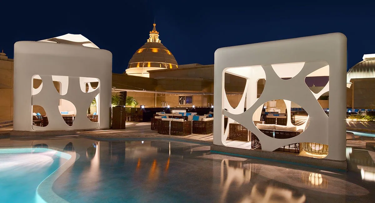 Night view of an outdoor pool area with modern white screens featuring abstract cutouts, overlooking a domed building with golden lighting.
