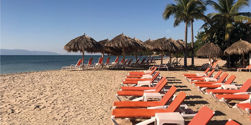 Empty orange lounge chairs and thatched umbrellas on a sandy beach with palm trees and calm water in the background.