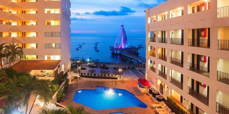 View of a hotel balcony overlooking a pool, with the ocean and a lit-up pier with a sailboat statue and boats in the background at dusk.