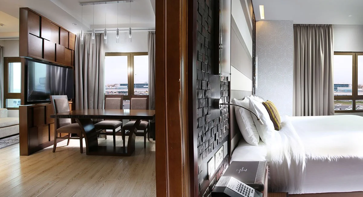Split view of a modern hotel suite with a dining area and a bedroom, separated by a wooden partition, featuring large windows with city views, curtains, and contemporary decor.