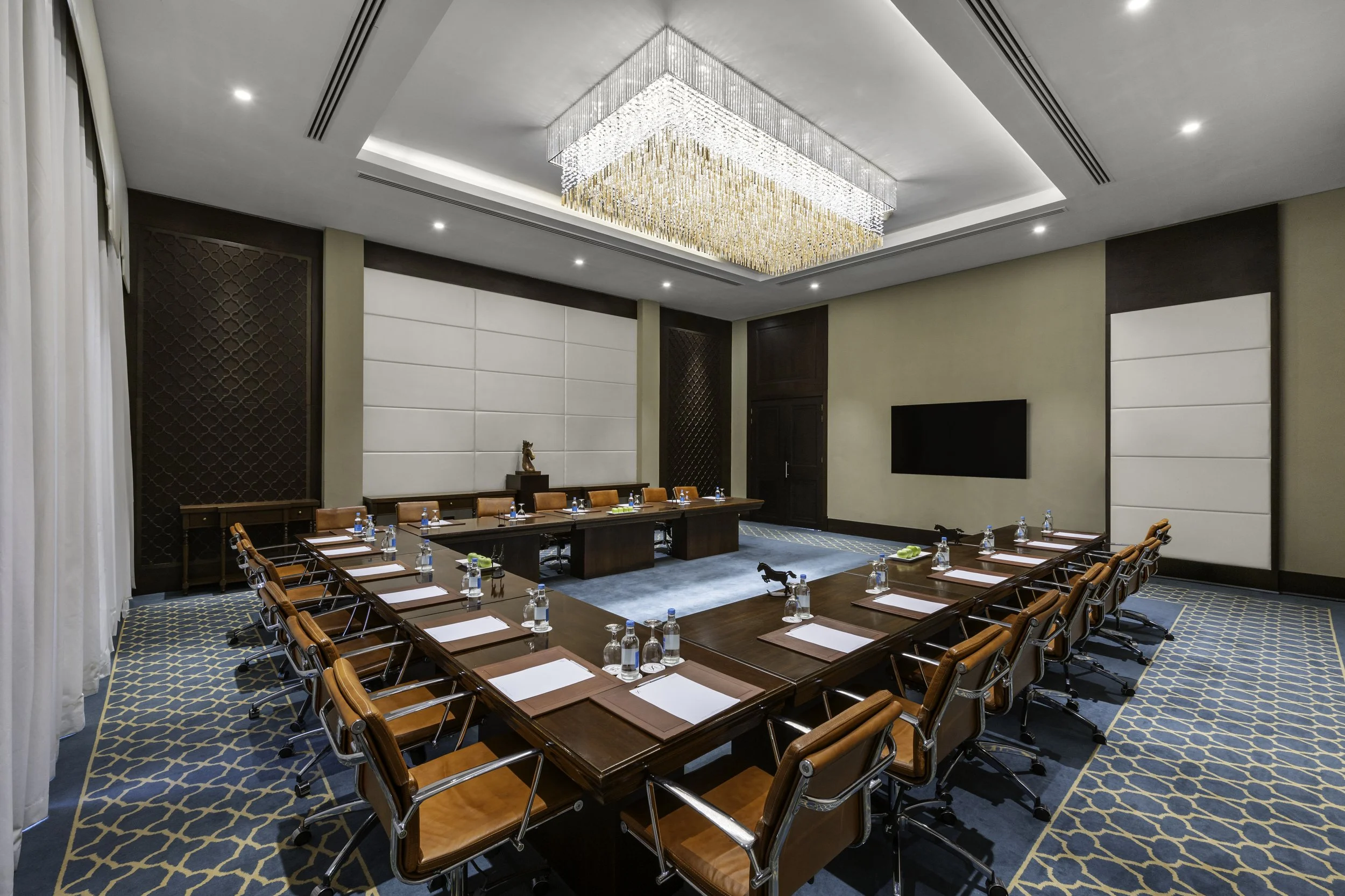 Conference room with a long U-shaped table set with notepads, bottled water, and pens, surrounded by wooden chairs with leather seats, and a large crystal chandelier hanging from the ceiling.