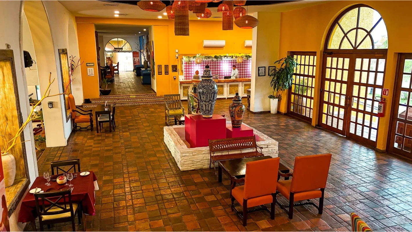 Hotel lobby with terracotta tile flooring, yellow walls, and wooden accents. Central display of decorative vases on a white brick platform. Seating areas include orange and upholstered chairs, some with small tables. Large arched wooden doors and win