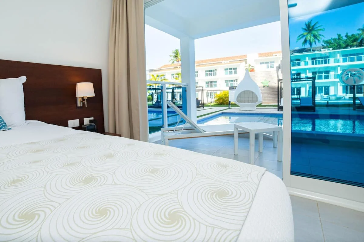 Interior of a bedroom with a bed, bedside lamp, and large sliding glass door leading to a balcony with a view of a swimming pool and modern buildings.