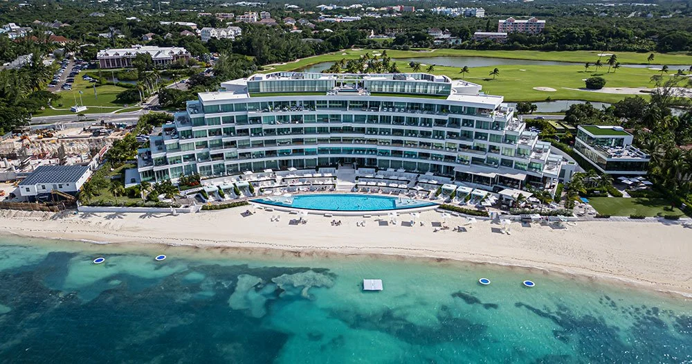 Aerial view of a modern beachfront resort with a curved building, swimming pools, and white sandy beach near turquoise waters, with green fields and trees in the background.