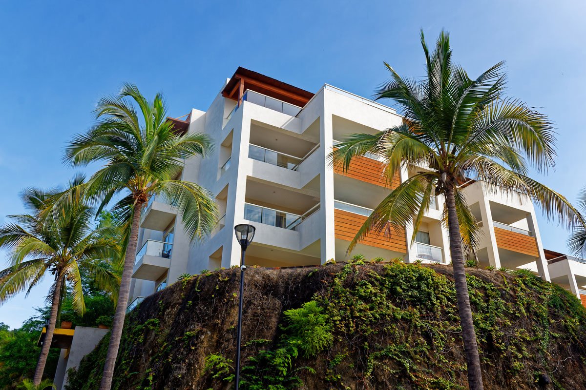 Modern white apartment building on a hillside surrounded by palm trees under a clear blue sky.