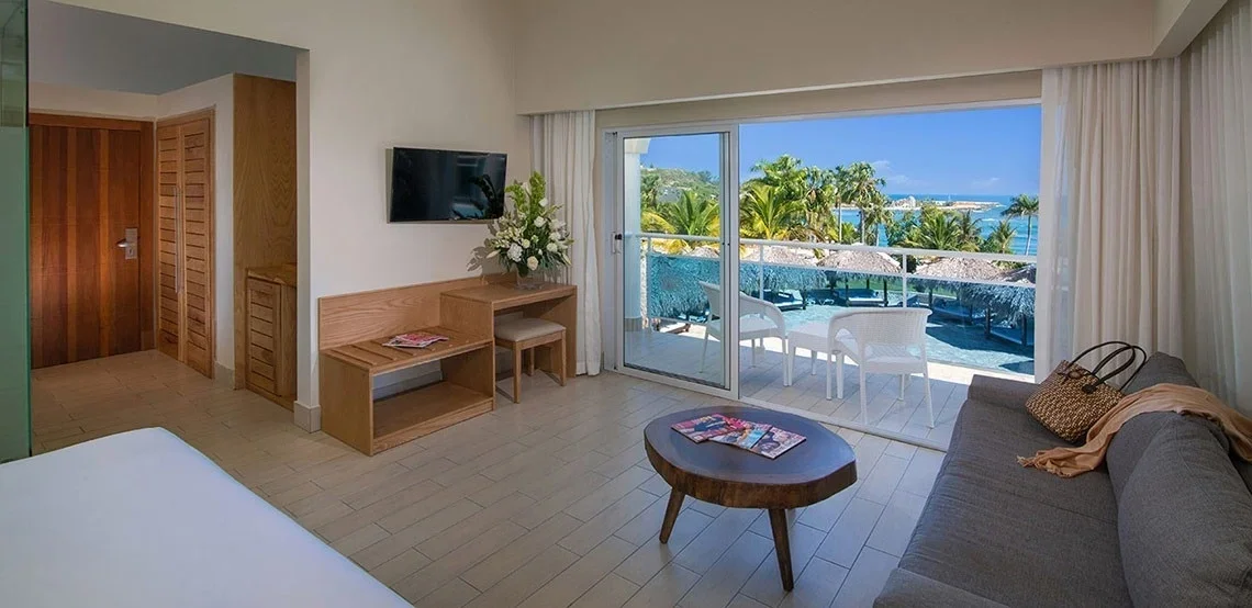 Beachside hotel room with sliding glass door leading to balcony with ocean view, white chairs, tropical palm trees, gray sofa with pillows, wooden coffee table, flat-screen TV, and wooden closet.