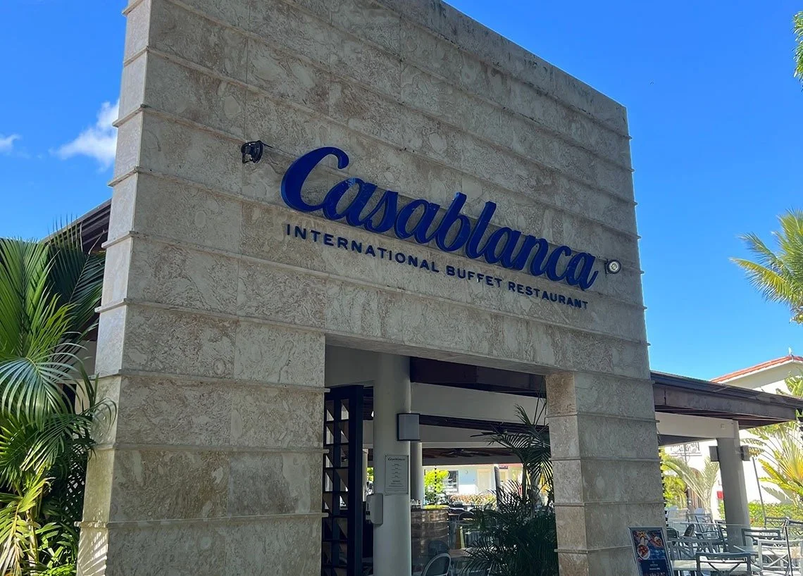 Entrance of Casabella, an international buffet restaurant with a beige stone facade, blue signage, and tropical landscaping.