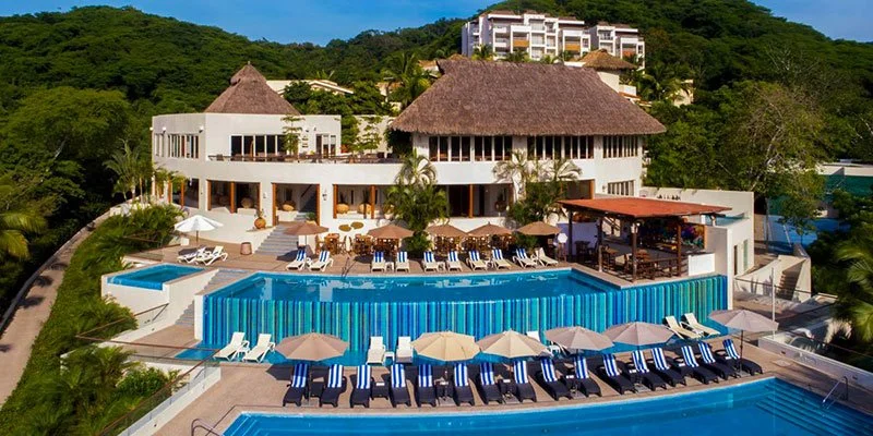 Luxury resort with a swimming pool, lounge chairs with umbrellas, and white buildings with thatched roofs surrounded by lush green trees.
