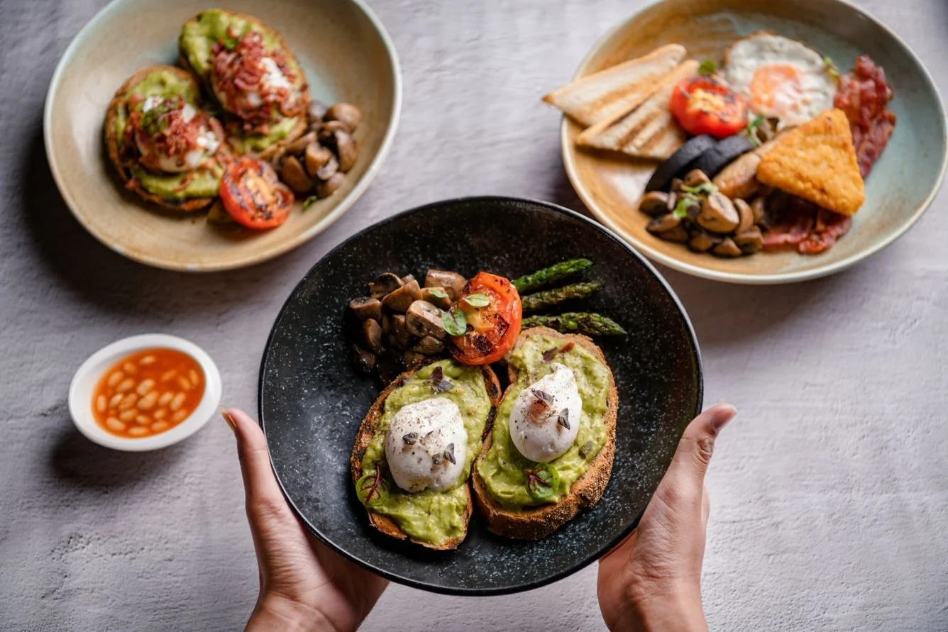 A person holding a black plate with avocado toast topped with poached eggs and garnished with herbs, along with roasted vegetables including cherry tomato, asparagus, and mushrooms. In the background, there are two beige plates with more food, includ