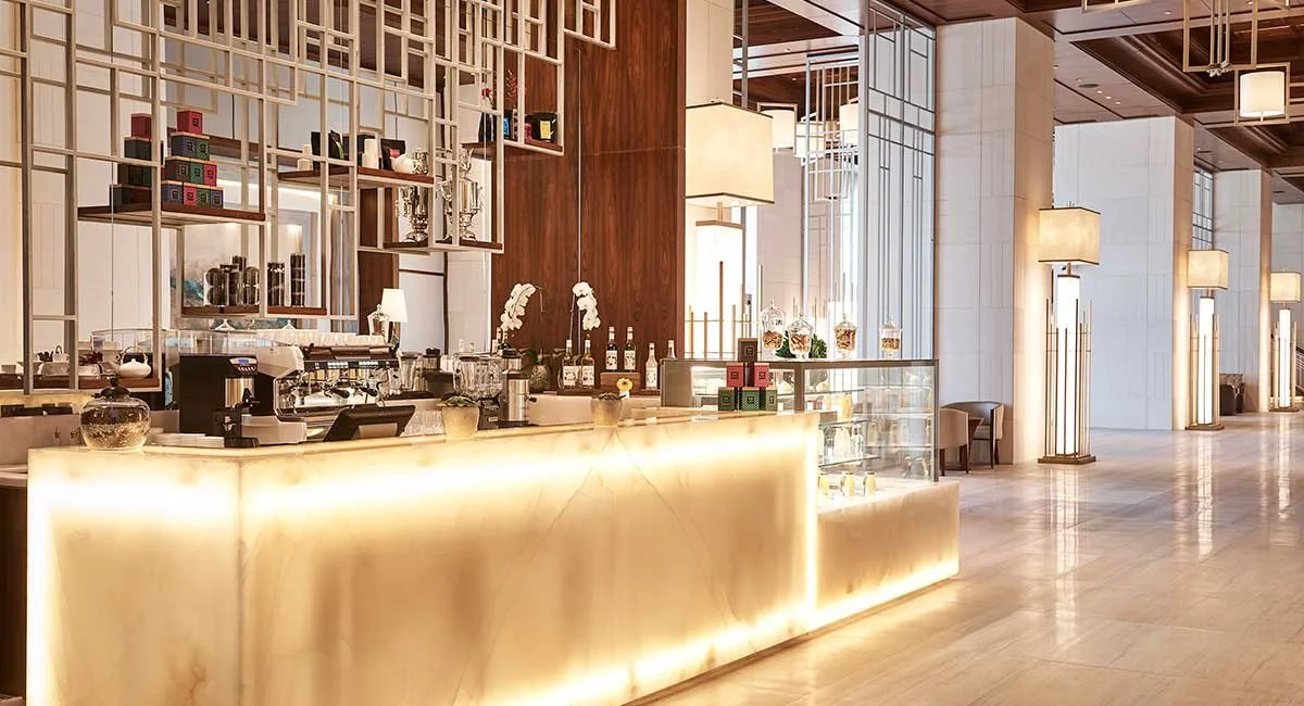 Luminescent hotel reception counter with decorative jars, plants, and a coffee machine, illuminated by warm lighting, with seating and large windows in the background.
