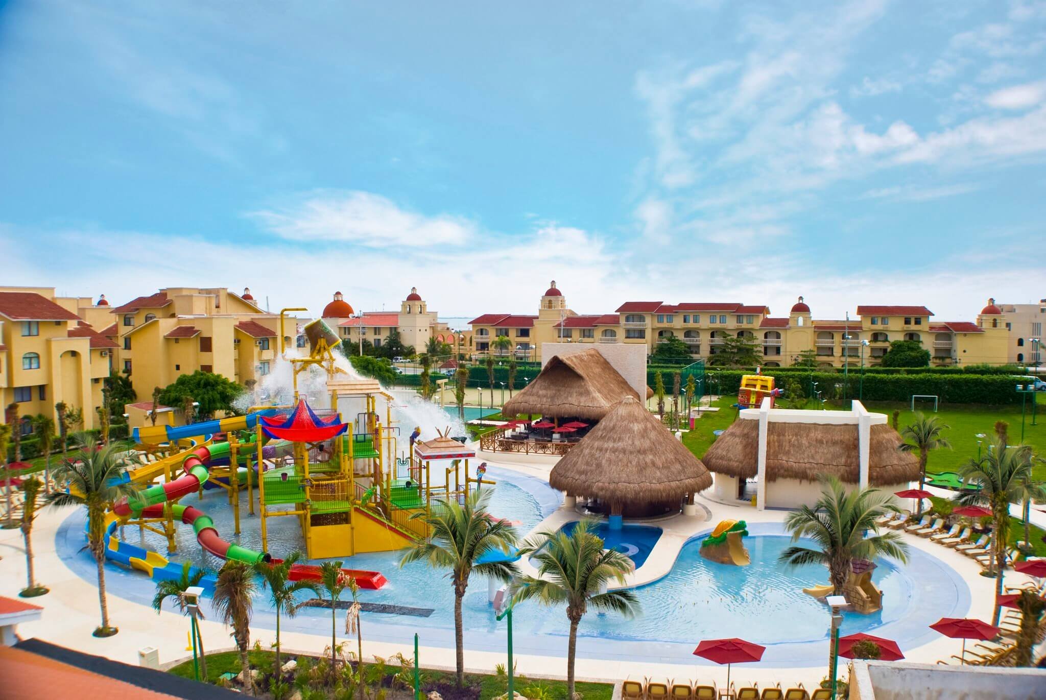 View of a colorful water park with water slides, lazy river, thatched roof structures, umbrellas, and palm trees, with residential buildings and blue sky in the background.