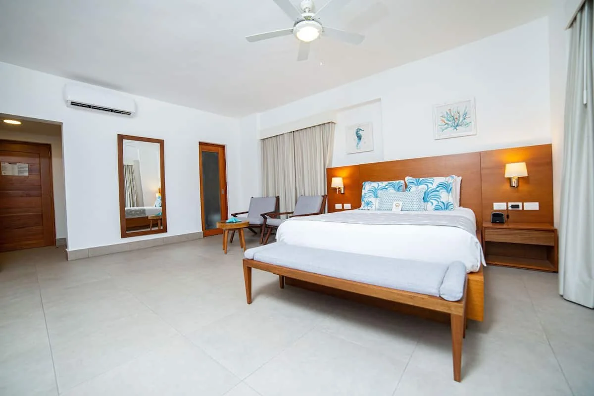 An hotel room with a large bed, wooden headboard, two pillows with blue and white patterns, two bedside lamps, a sitting area with two chairs and a small table, a mirror, and a door, with curtains and ceiling fan.