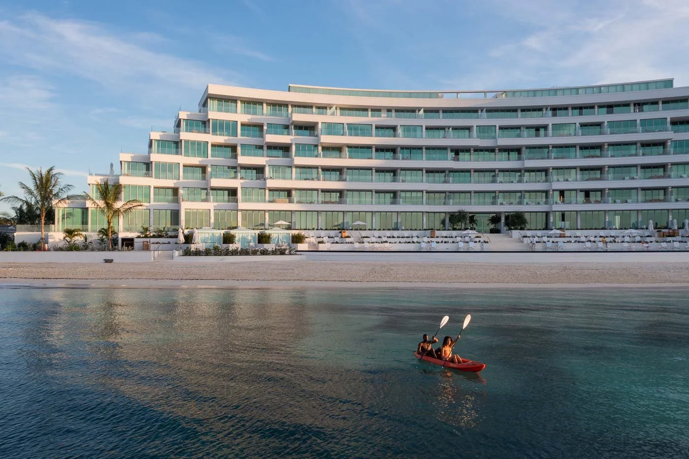 Two people kayaking in front of a large modern beachfront hotel with multiple glass balconies, palm trees, and lounge chairs.