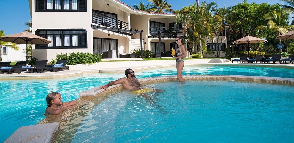 A family enjoying a swim in a resort pool near a white multi-story building with balconies and tropical palm trees, under a sunny sky with umbrellas and lounge chairs around the pool.