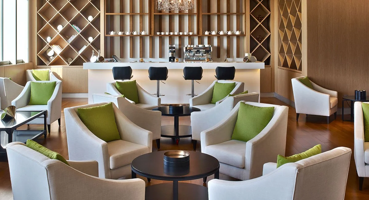 A stylish lounge with white armchairs and green cushions arranged around small black tables, a bar with four black barstools behind it, wooden wall decor, and a coffee machine on the counter.