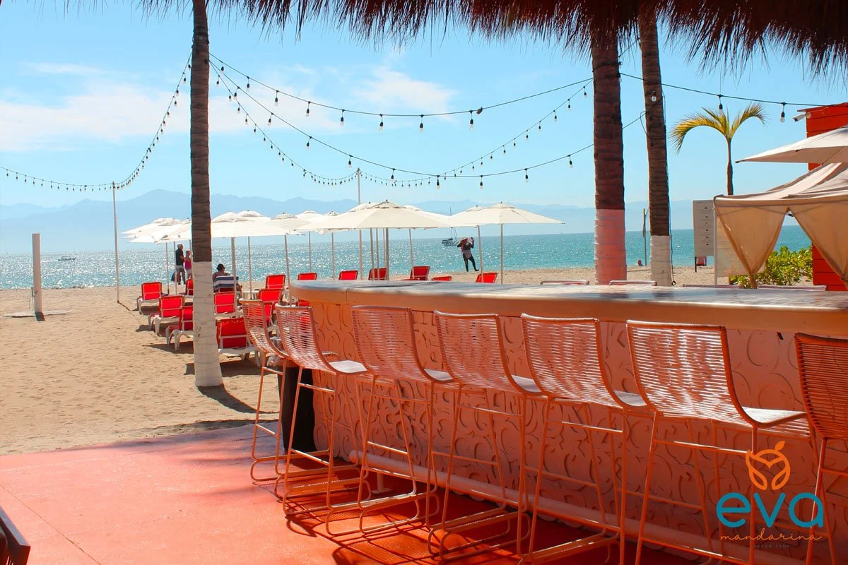 View of a beach bar with a long counter and orange bar stools, overlooking the sandy beach. There are palm trees, string lights, and white umbrellas on the beach, with the ocean and mountains in the background under a blue sky.