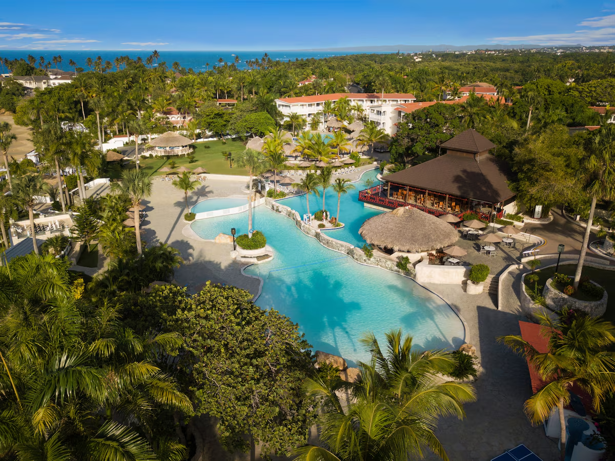 An aerial view of a tropical resort with a large, winding swimming pool surrounded by lush palm trees, a thatched-roof hut, and a large open-air restaurant or bar with a dark roof. The resort is located near the ocean, visible in the background, with