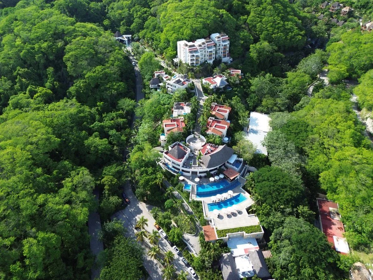 Aerial view of a large luxury house complex surrounded by dense green forest with multiple swimming pools and outdoor lounging areas.