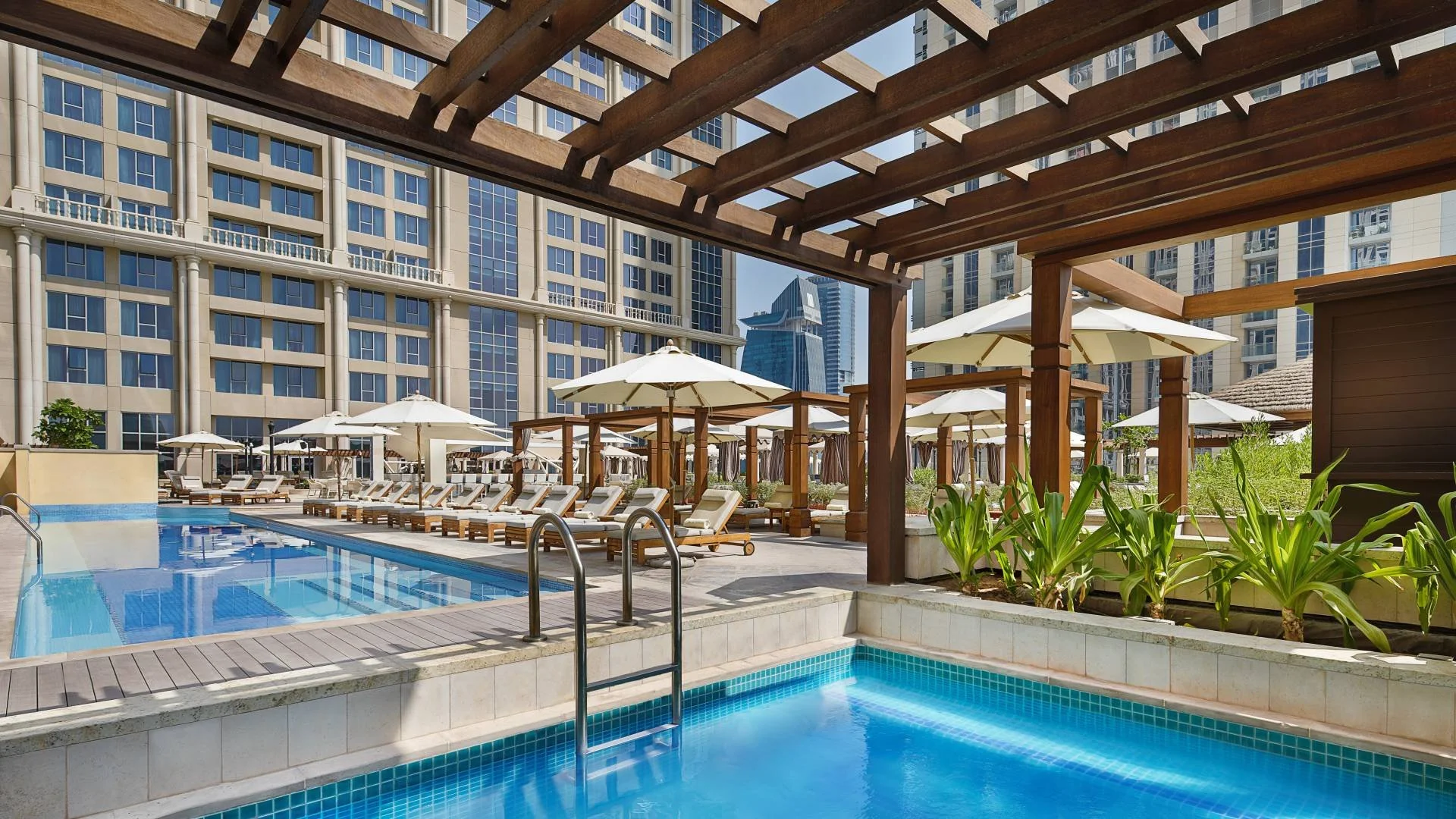 Indoor swimming pool area with blue-tiled pool and hot tub, poolside lounge chairs with umbrellas, wooden pergolas, and tall modern buildings in the background.