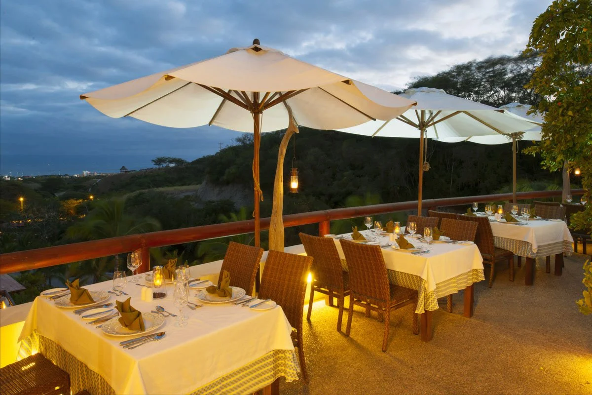 An outdoor restaurant patio at dusk with tables set with white tablecloths, place settings, and lit candles, shaded by large white umbrellas, overlooking a hilly landscape with trees and a cloudy sky.