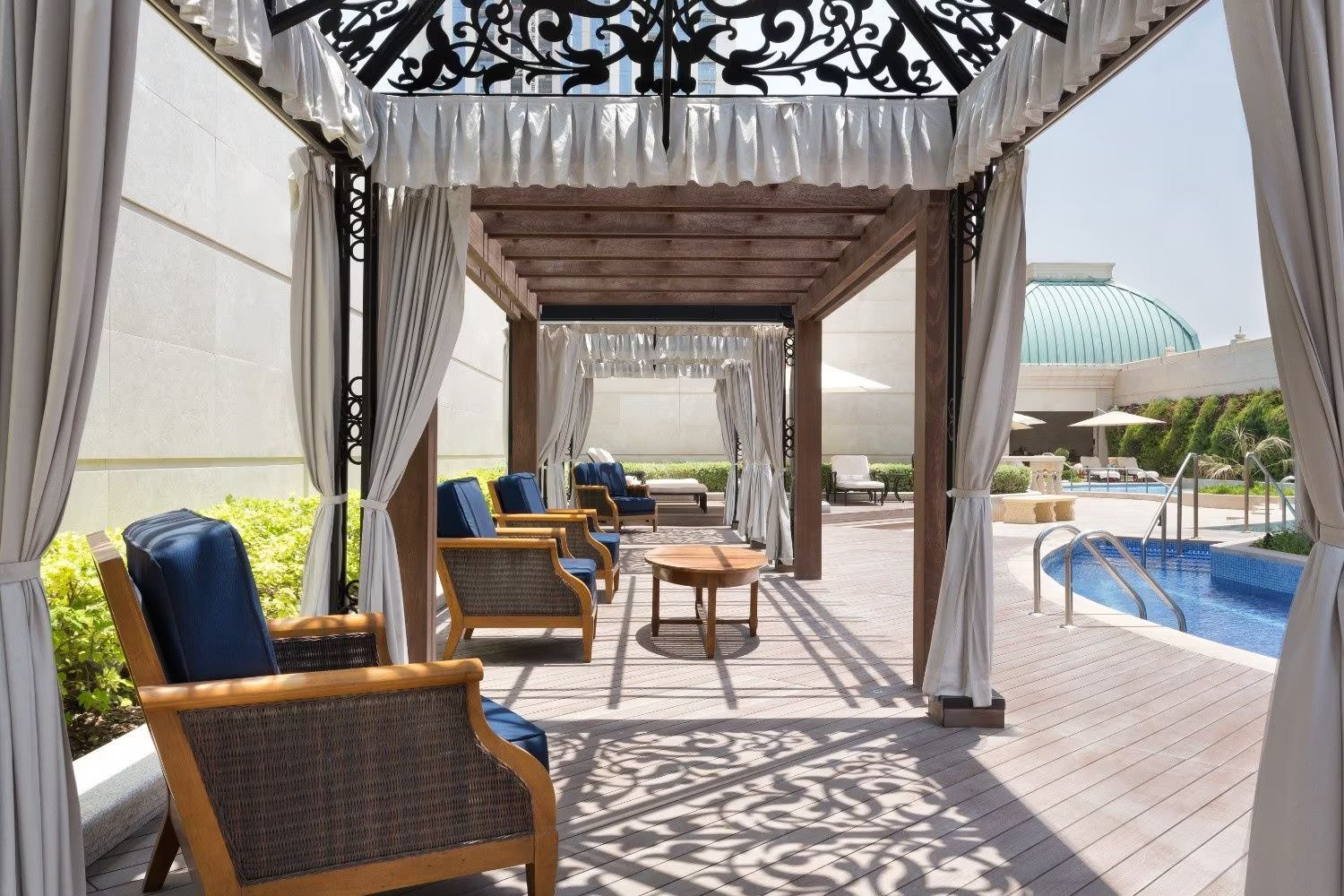 Outdoor poolside cabanas with blue cushioned chairs, wooden tables, and white curtains, overlooking a swimming pool with a building in the background.