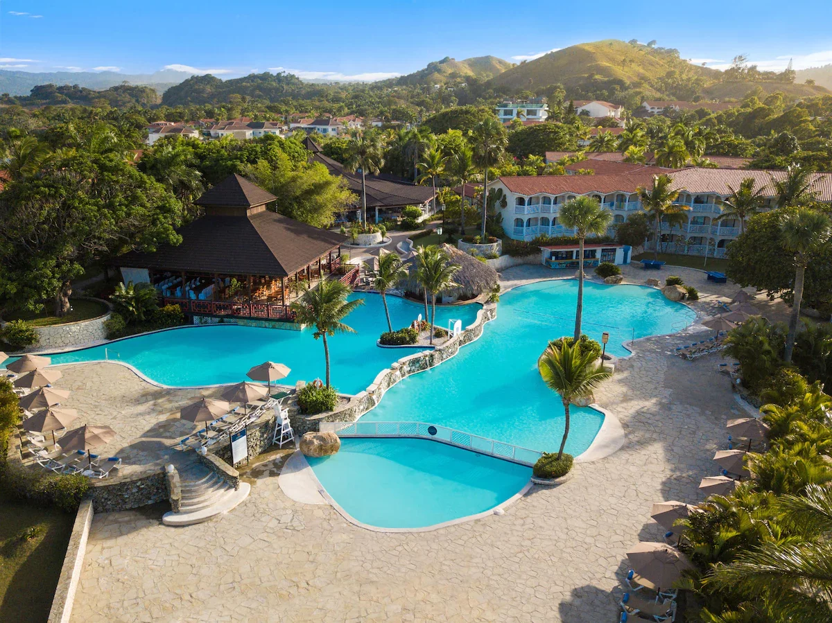 Aerial view of a resort pool area surrounded by lounge chairs, umbrellas, palm trees, lush greenery, and buildings with hills in the background.