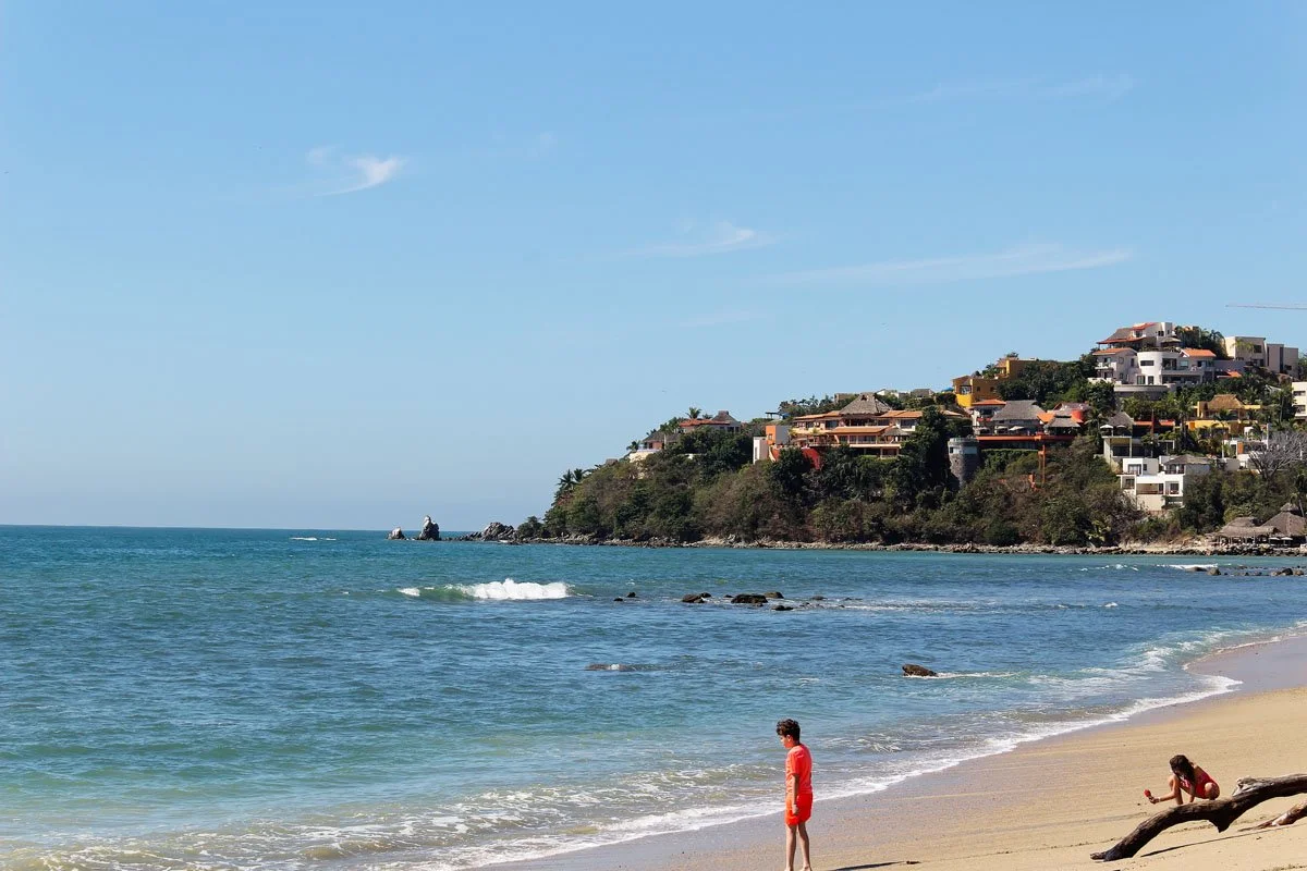 People on a sandy beach near the ocean with houses on a hill in the background.