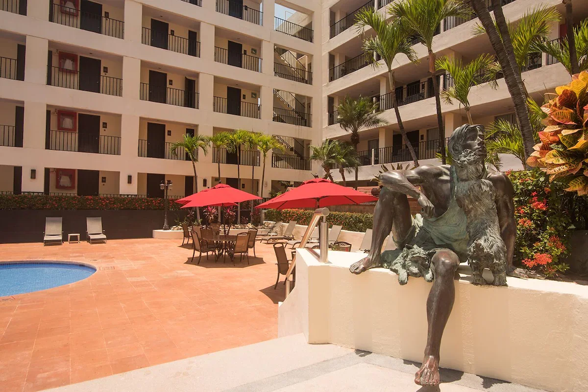 Courtyard of a hotel with a swimming pool, outdoor chairs with red umbrellas, palm trees, and a statue of a bearded man with one leg over a dog, in front of a white multi-story building.