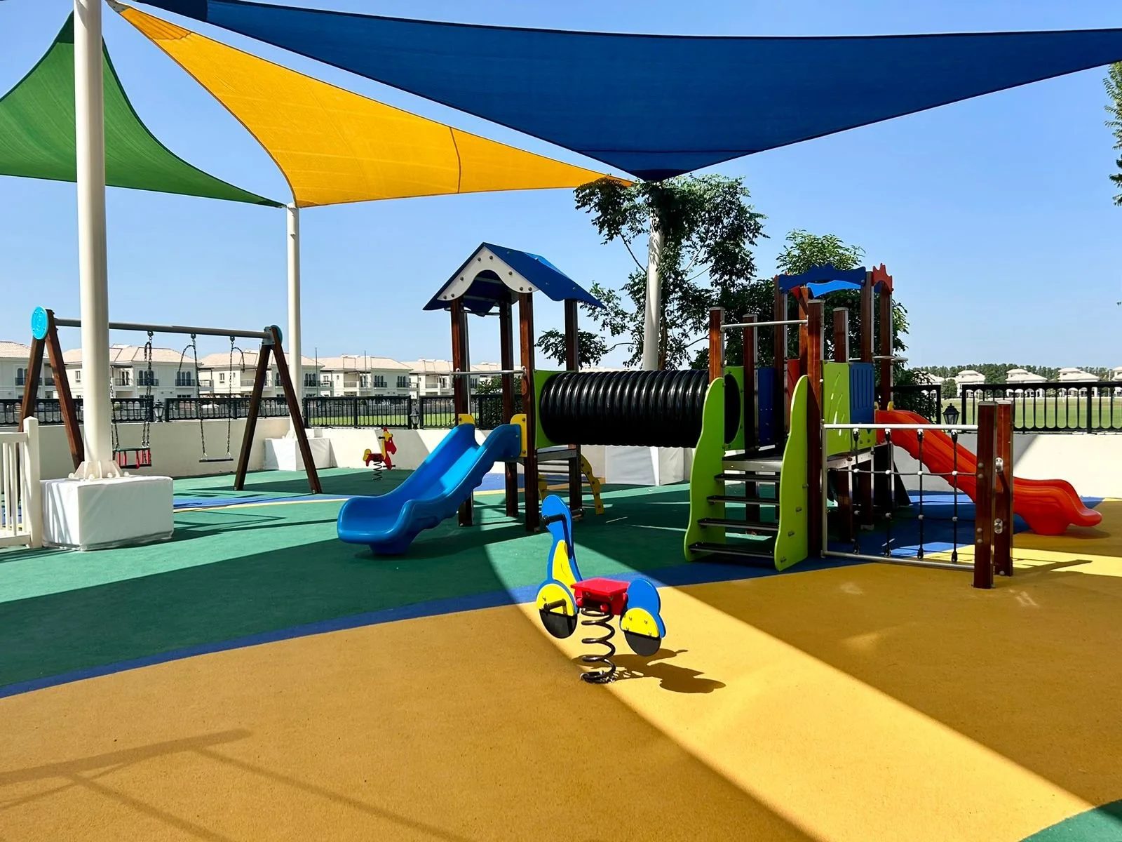 Colorful outdoor playground with slides, a tunnel, swings, and spring riders under a large shade sail.