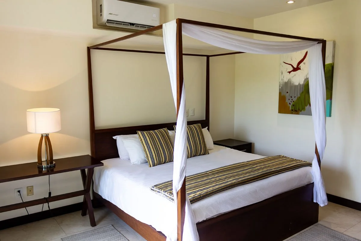 A bedroom with a four-poster bed featuring a wooden frame, white curtains, and striped pillows and bed runner. There is a bedside table with a lamp on the left side, a small bedside table on the right, a colorful painting on the wall, and an air cond