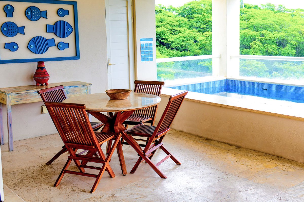 A sunlit outdoor patio with a round wooden dining table and four matching wooden chairs. There is a bowl on the table, a colorful red lamp on a distressed wooden console table, and a blue fish-themed wall art above the console. Large windows reveal l
