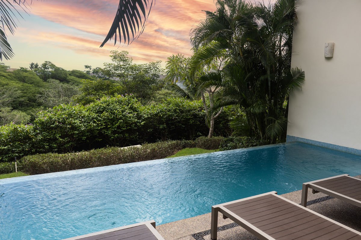 Infinity pool overlooking a lush green landscape and colorful sunset sky, with palm trees and tropical plants nearby.