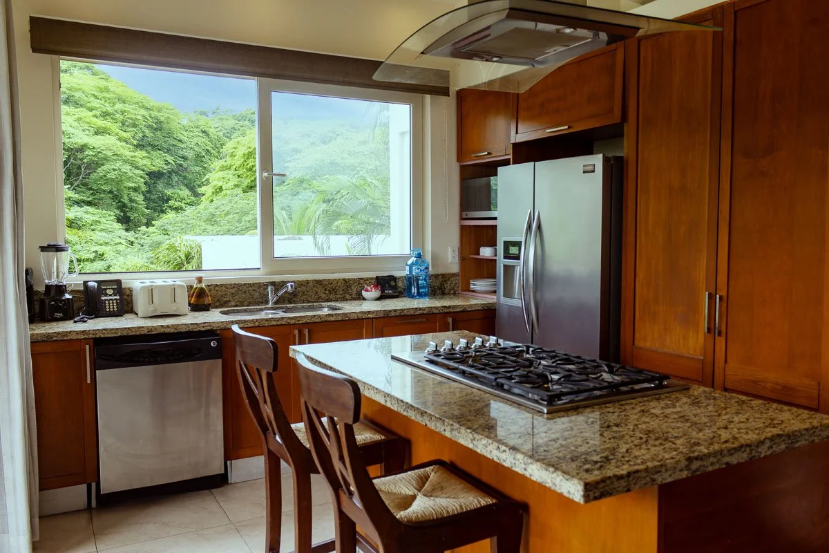 Kitchen with wooden cabinets, granite countertops, stainless steel refrigerator, gas stove, microwave, blender, toaster, water bottle, and a window showing lush green trees outside.