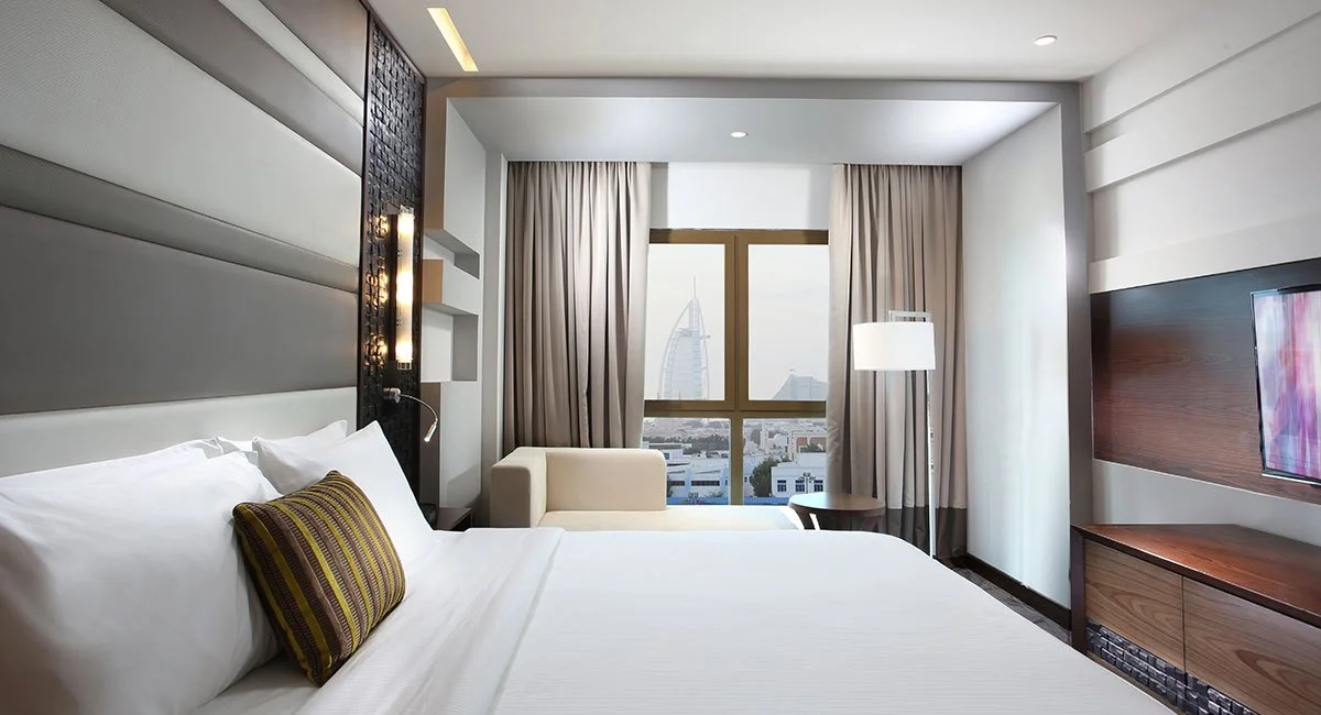 A hotel room with a large bed, white linens, and a decorative pillow, facing a window with a view of the city, including the Burj Al Arab hotel.
