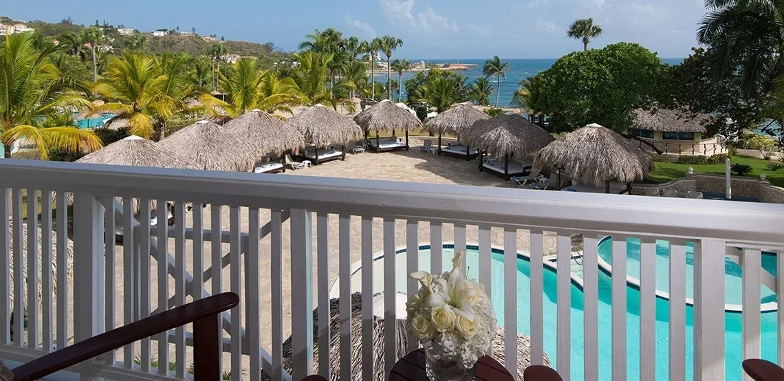 View from a balcony overlooking a pool area with straw-roofed cabanas, tropical trees, and the ocean in the background.