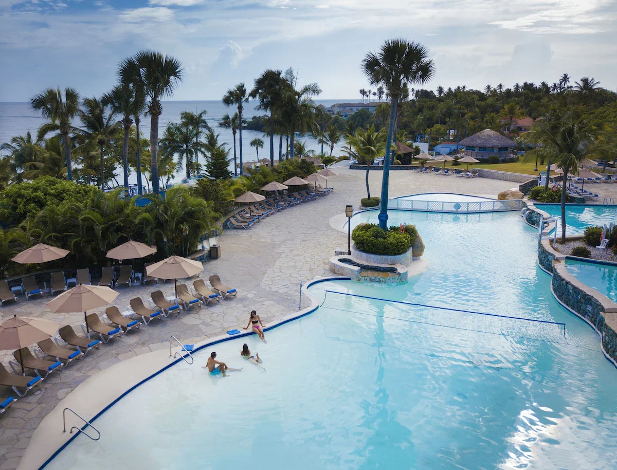 Beachside resort with swimming pool, lounge chairs with umbrellas, palm trees, and ocean in the background.