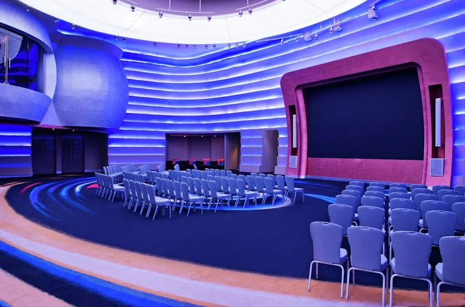 An empty modern auditorium with a large screen, theater-style seating, and ambient blue lighting.