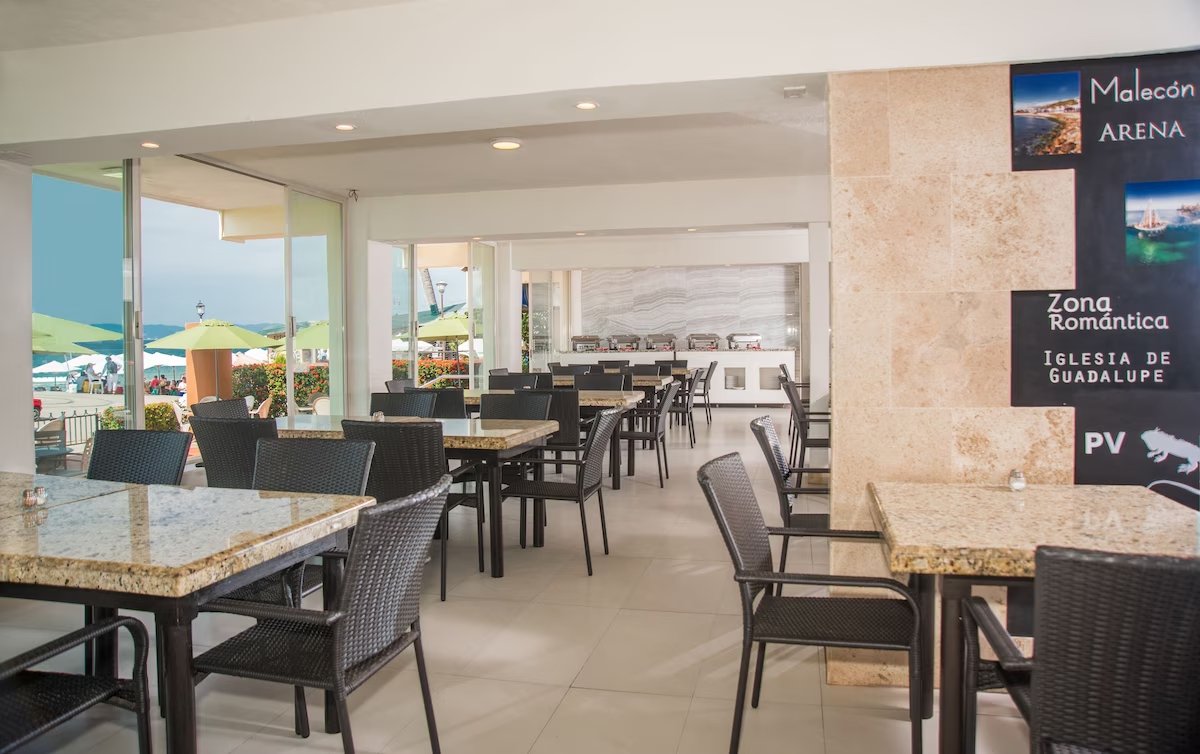 Indoor restaurant area with granite-topped tables and black wicker chairs. Outside, green umbrellas shade outdoor seating with a view of the ocean and beach. A sign on the wall indicates locations including Malecon Arena, Zona Romantica, and Iglesia 