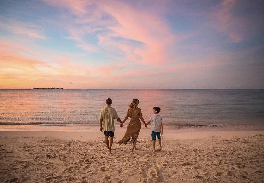 A family of three holding hands while walking on the beach during sunset, with pink and purple clouds in the sky and calm ocean water.