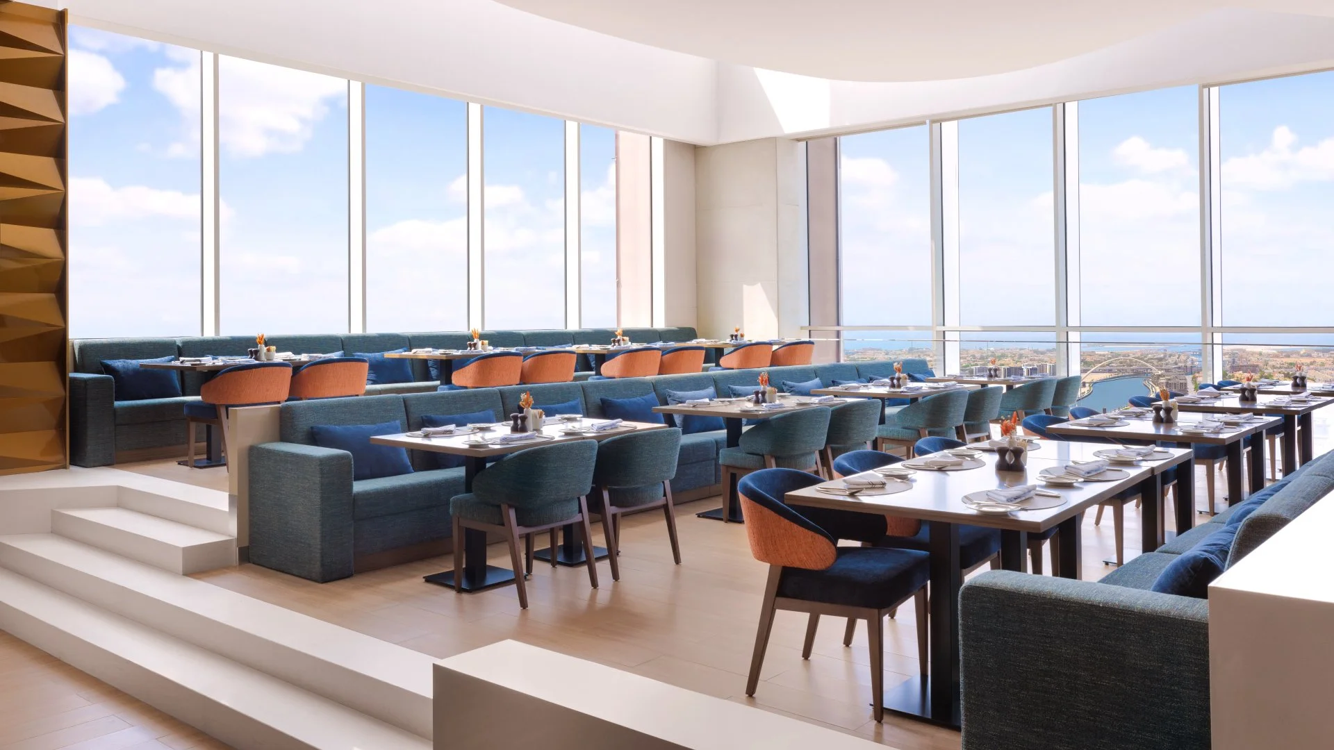 Upscale restaurant with large floor-to-ceiling windows offering panoramic city views. Stylish tables set with white plates, silverware, and small flower arrangements. Contemporary blue and peach upholstered chairs and cushioned banquettes along the w