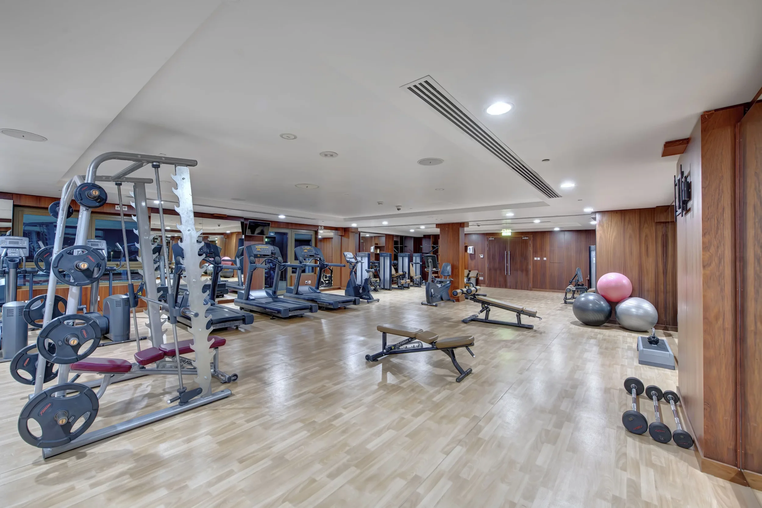A spacious gym with various workout equipment including weight plates, treadmills, exercise bikes, and stability balls, with wooden paneling on the walls and a white ceiling with recessed lighting.
